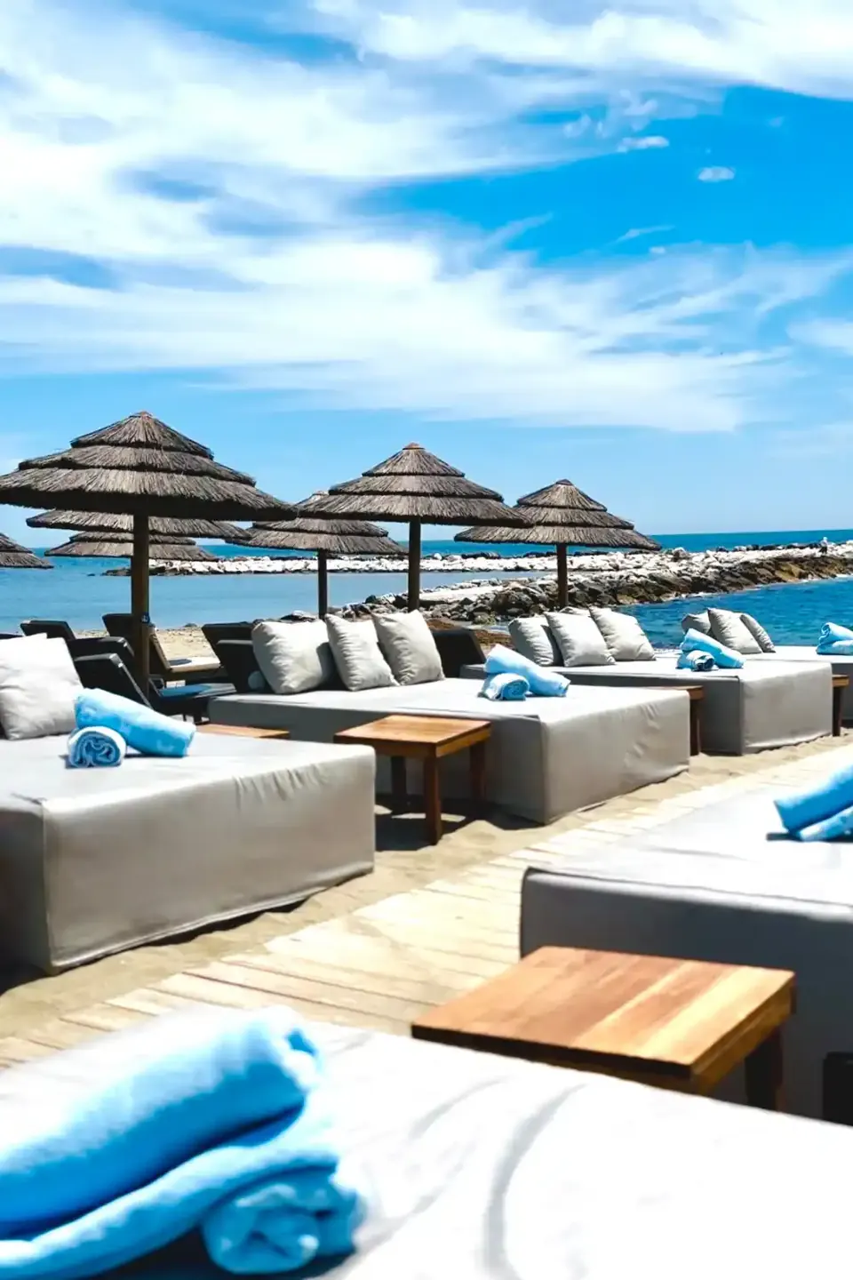 Camas de playa con cojines y toallas azules bajo sombrillas de brezo frente al mar